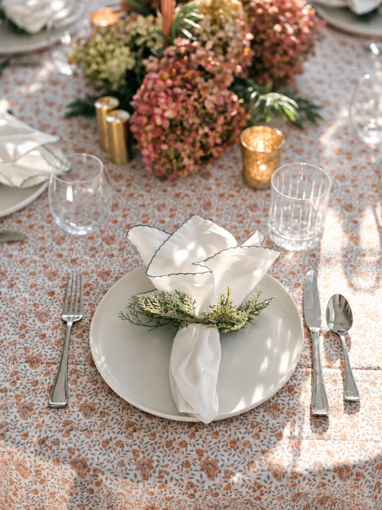 Ivory plate with a white linen napkin tied with greenery on a floral tablecloth, with hydrangeas and gold votives.