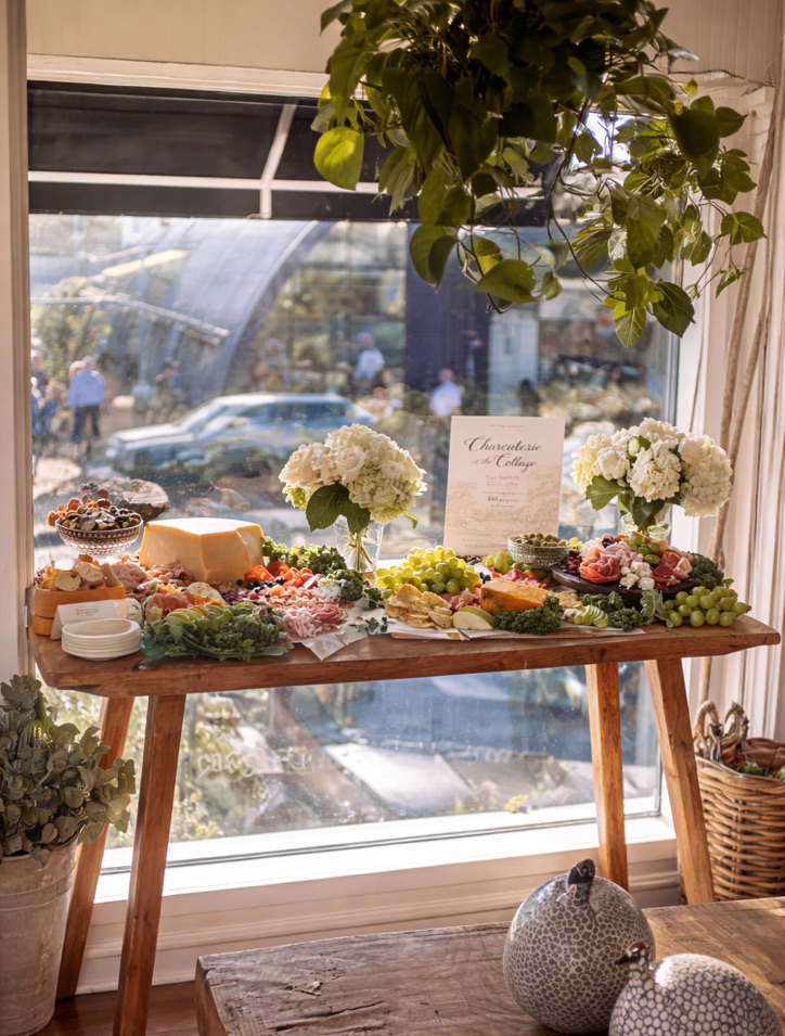 A sunlit charcuterie spread featuring cheeses, cured meats, fresh fruit, and savory bites, styled with white flowers on a rustic wooden table in Grange Garden’s Historic Cottage.