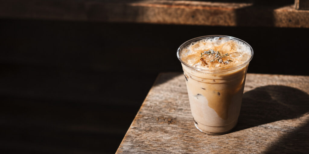 Creamy iced coffee catches the sunlight on a timeworn wooden table—a welcoming pause in your day at Grange Garden. Iced coffee in a clear cup on a vintage wooden table, illuminated by sunlight against a dark background.