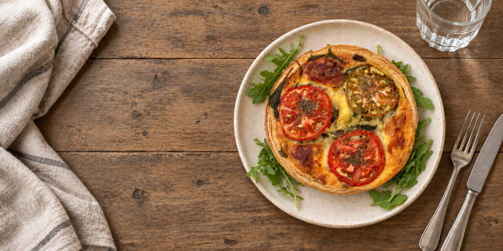 Our golden-crusted quiche, prepared by our private French Chef, Christophe Leneouanic, topped with roasted tomato slices and herbs, nestled on arugula and set on a rustic wooden table with soft linen.